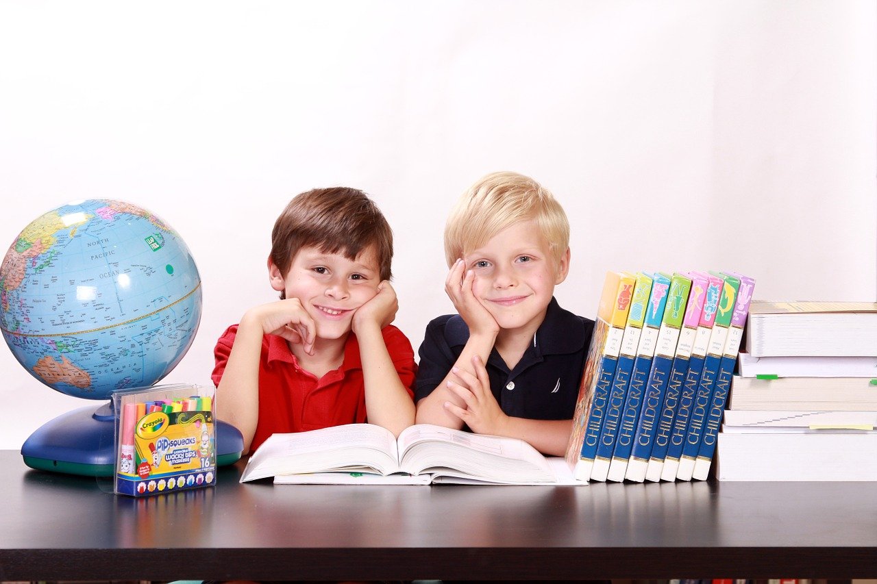dwóch chłopców siedzących przy biurku, na którym znajduje są globus, książki i przybory szkolne