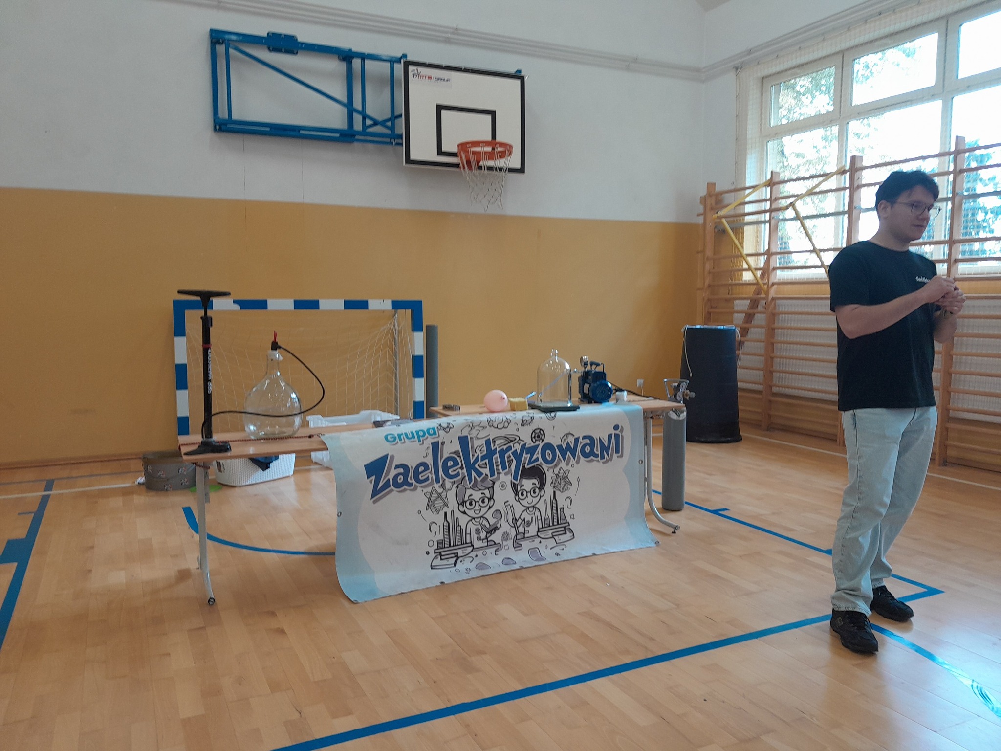 Przedstawiciel grupy "Zaelektryzowani" przy mikrofonie, za nim stolik z przyborami potrzebnymi do eksperymentów