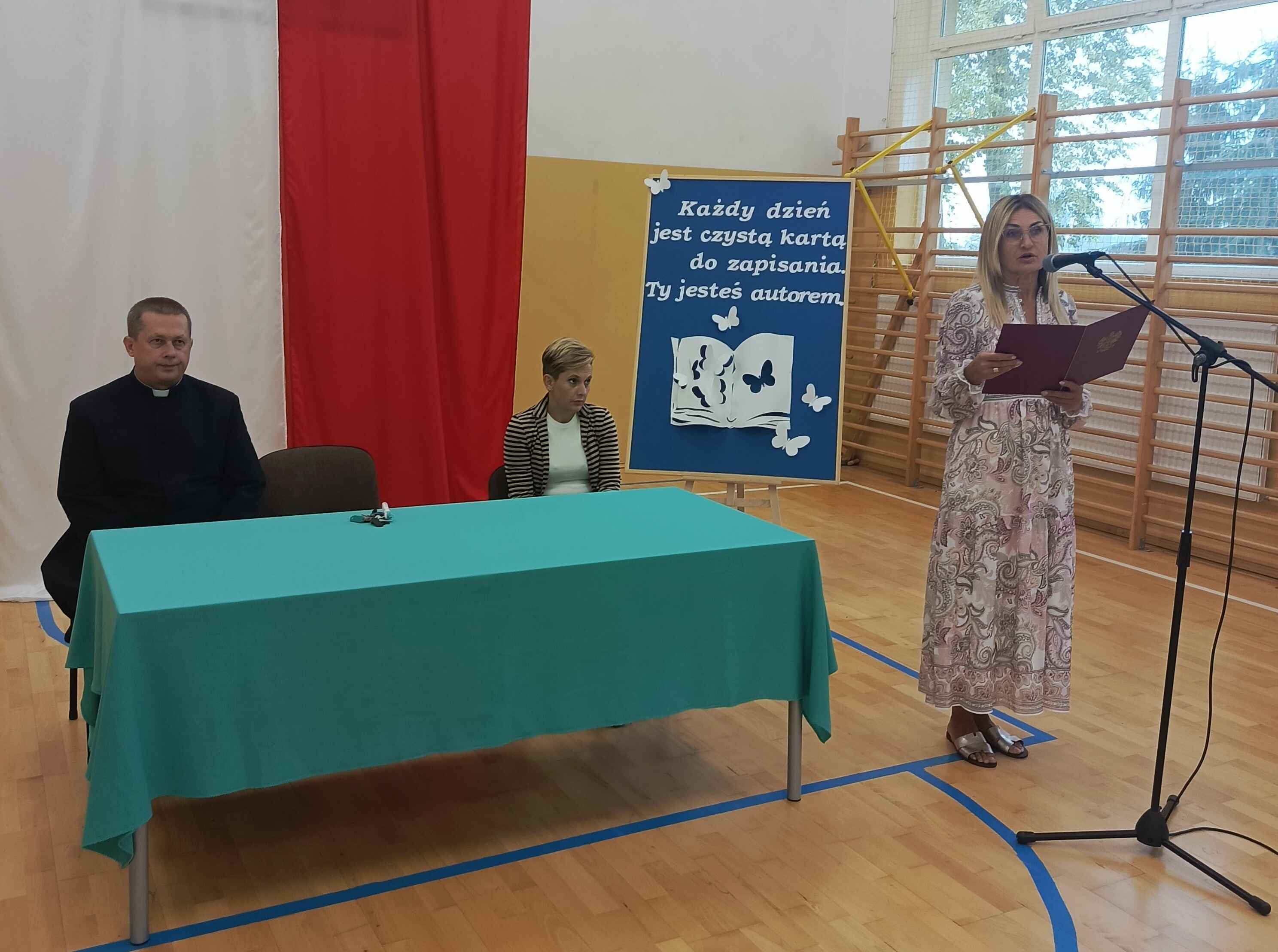Pani dyrektor przemawia przy mikrofonie na tle tablicy dekoracyjnej, obok przy stole przewodnicząca Rady Rodziców i ksiądz proboszcz parafii w Czerniczynie