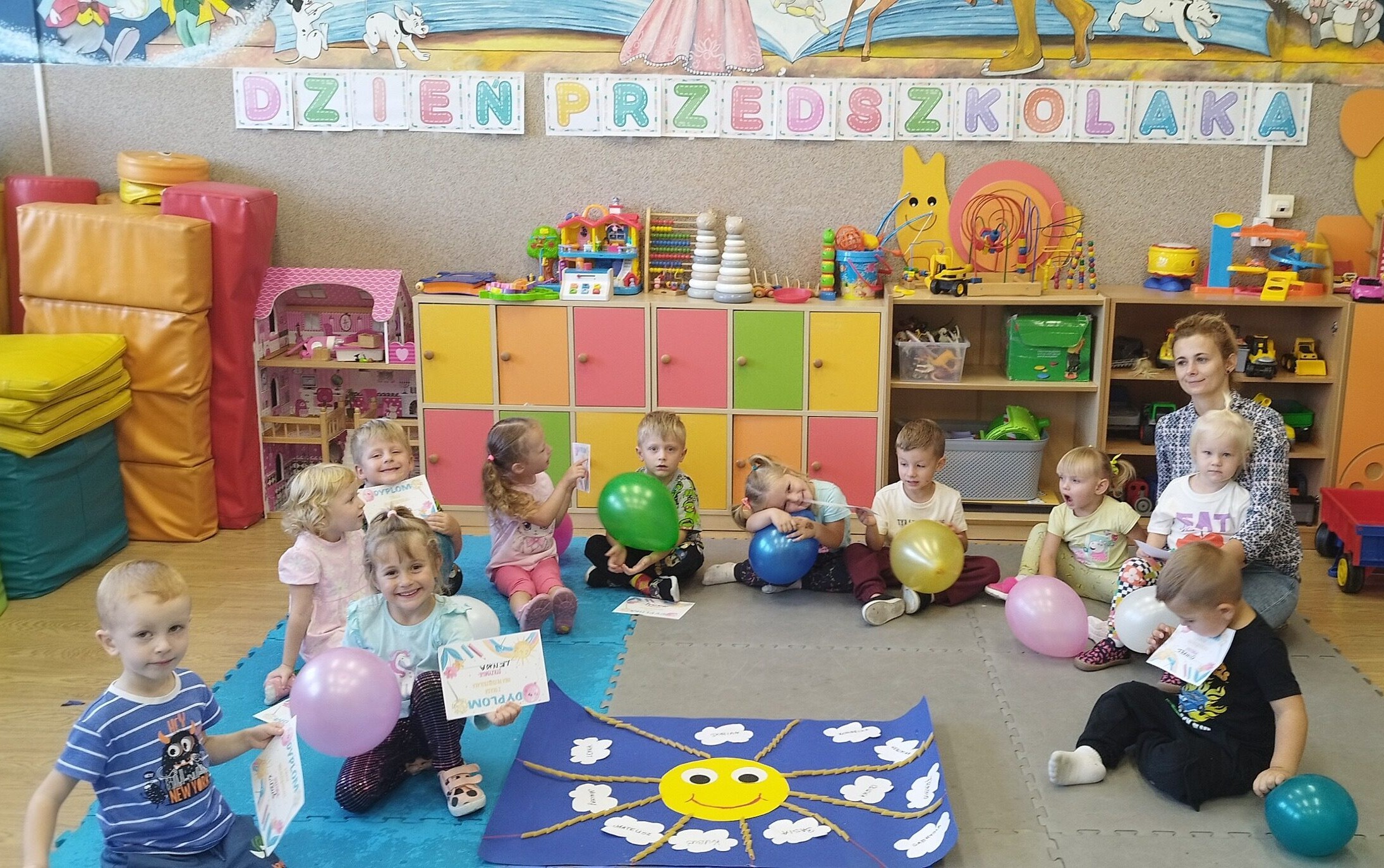 Przedszkolaki z wychowawczynią siedzą na dywanie wokół wspólnie wykonanego plakatu