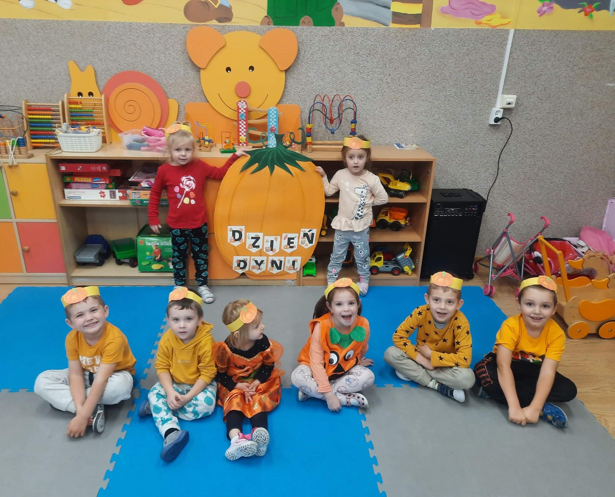 Przedszkolaki w pomarańczowych strojach i opaskach na głowach siedzą na dywanie na tle dyni z papieru