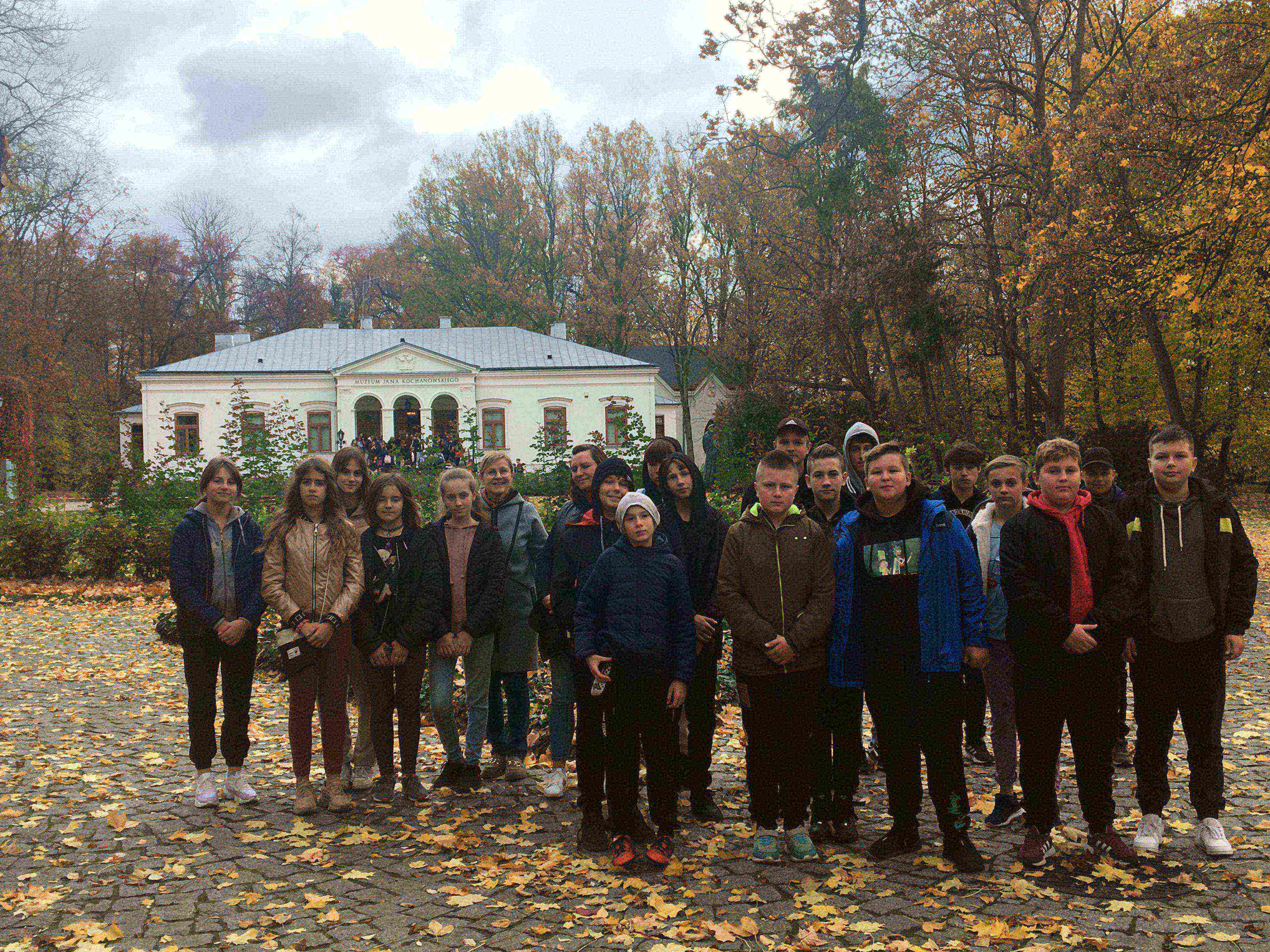 Grupa młodzieży na tle dworku otoczonego parkiem w barwach jesieni.