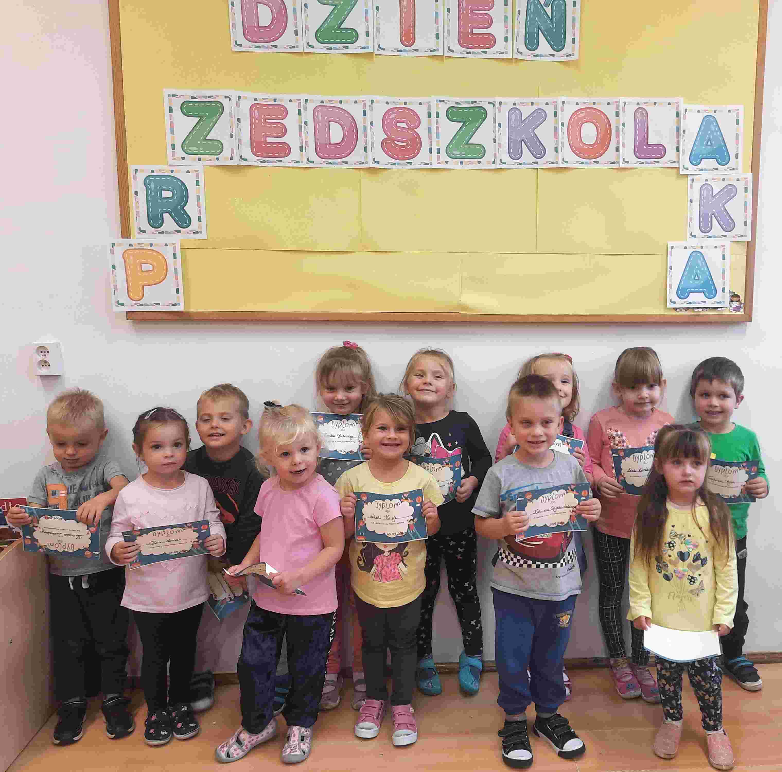 Grupa przedszkolaków z pamiątkowymi dyplomami pod tablicą z napisem: "Dzień Przedszkolaka"