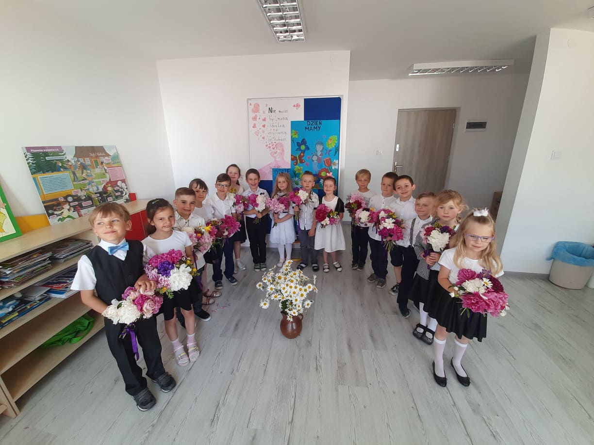 Dzieci w elegancich ubraniach trzymają bukiety kwiatów. Stoją półkolem wokół wazonu z polnymi kwiatami