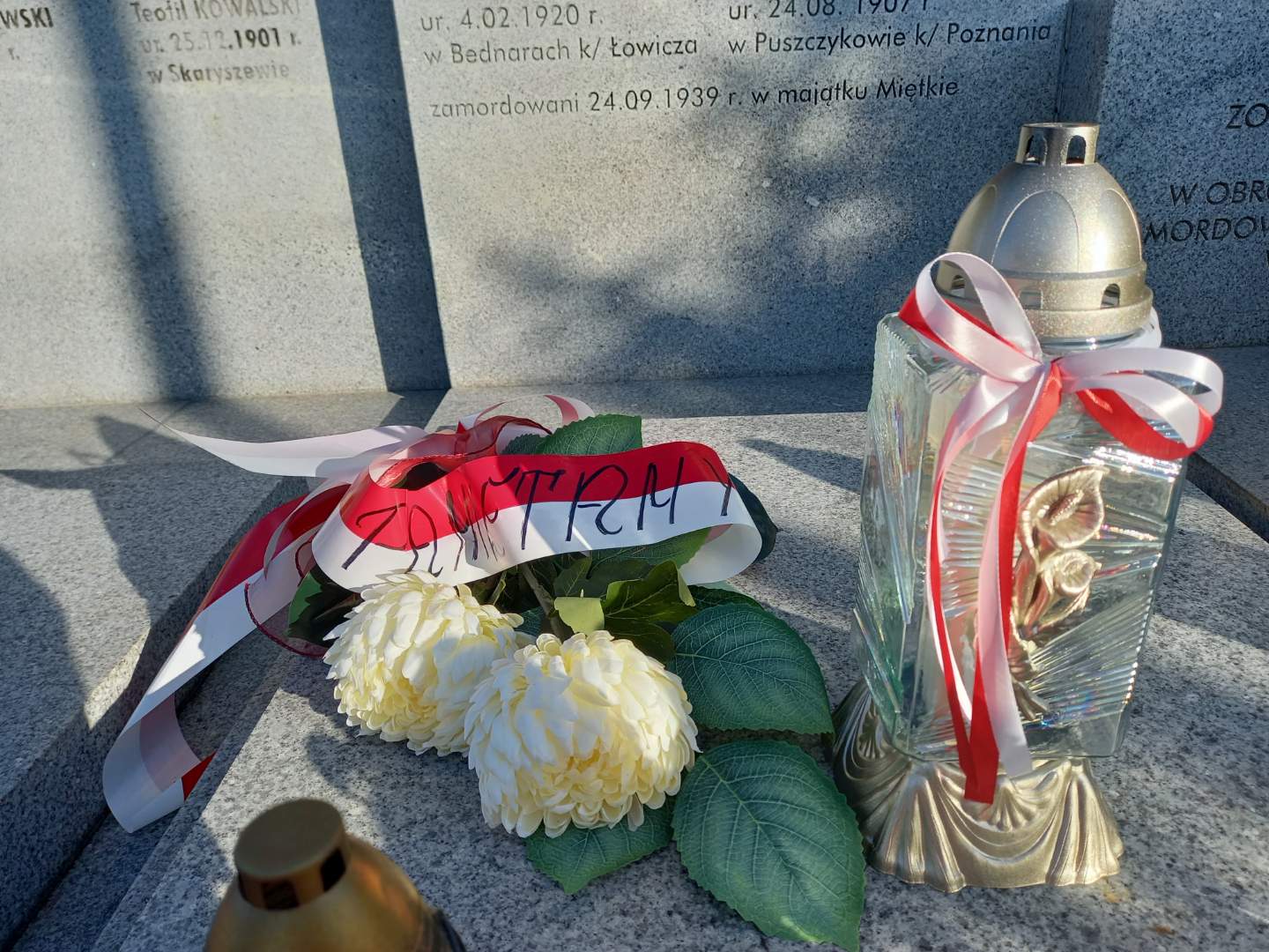 Zapalony znicz i kwiaty z biało-czerwonymi wstęgami w miejscu pamięci narodowej. Na wstędze napis: "Pamiętamy!"