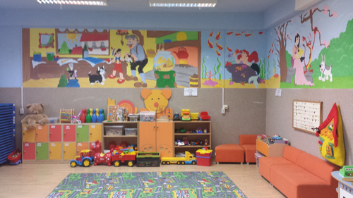 Pomieszczenie przedszkolne wyposażone w kolorowe szafki, zabawki, pomarańczową sofę oraz kolorowe obrazki na ścianach 