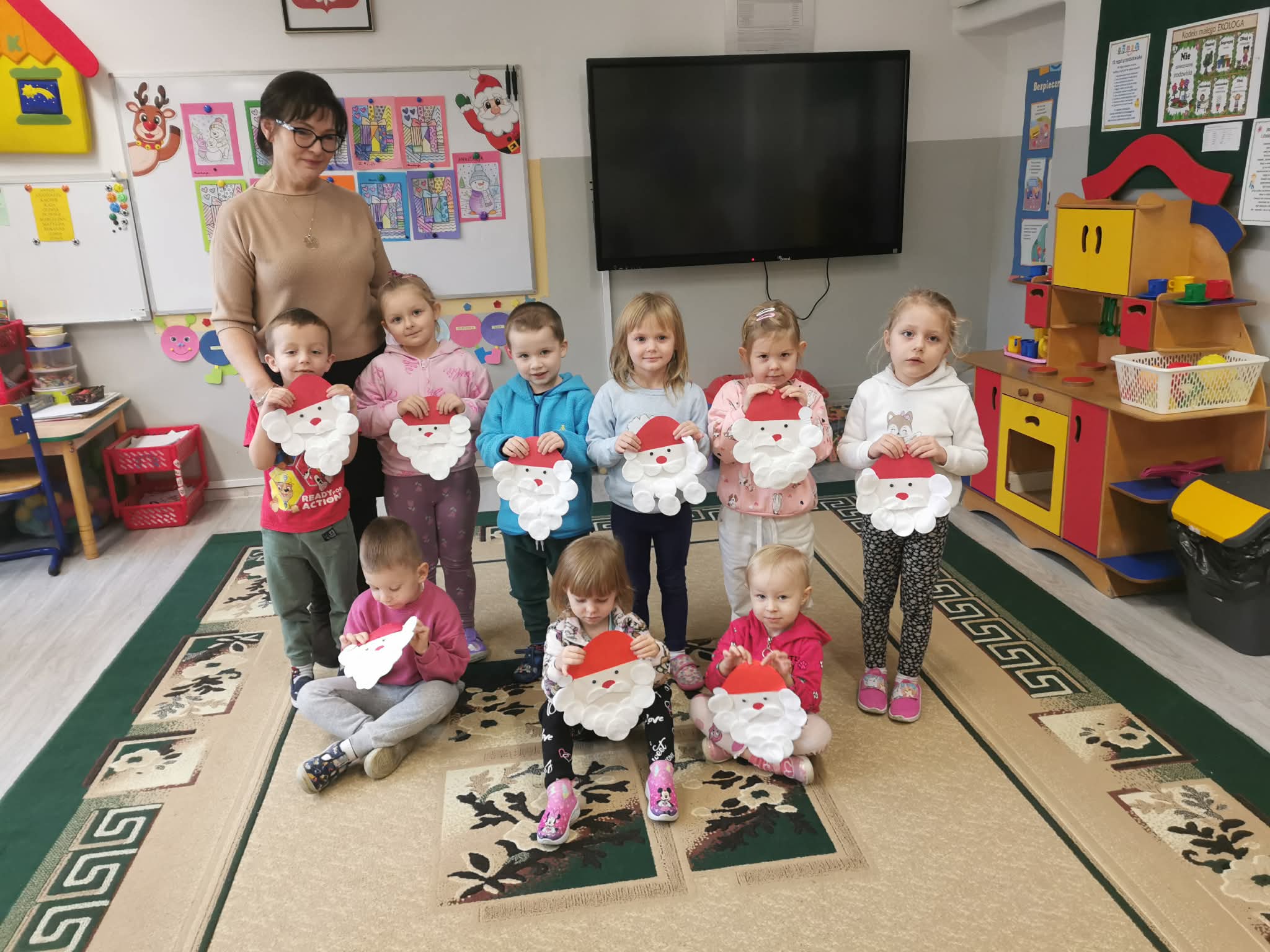 Grupa przedszkolaków pozująca z wyklejonymi portretami Mikołaja; obok stoi pani.