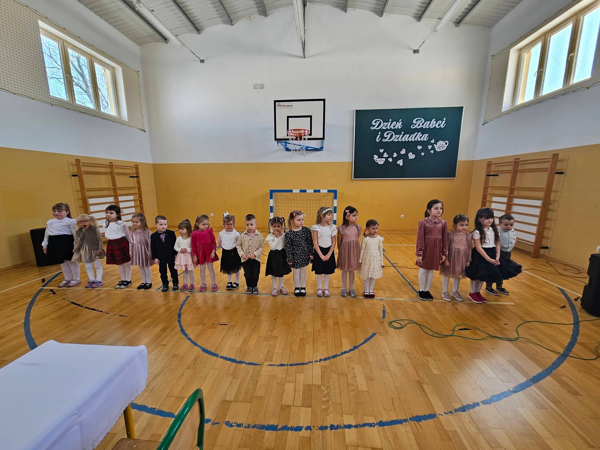 Dzieci w wieku przedszkolnym ustawione w szeregu na sali gimnastycznej, przygotowane do występu z okazji Dnia Babci i Dziadka.