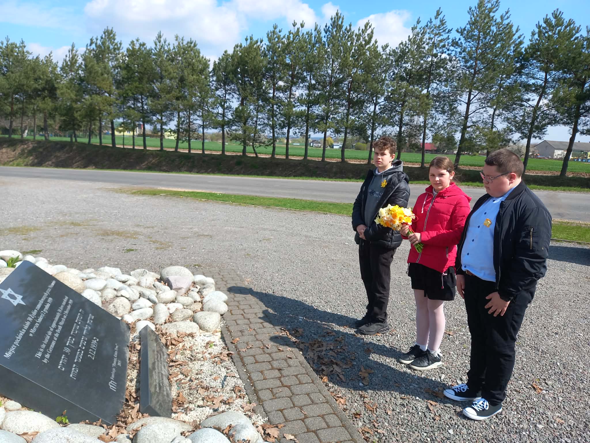 Uczniowie składają bukiet żółtych żonkili przy tablicy pamiątkowej w Cichoborzu, znajdującej się w otoczeniu kamieni.