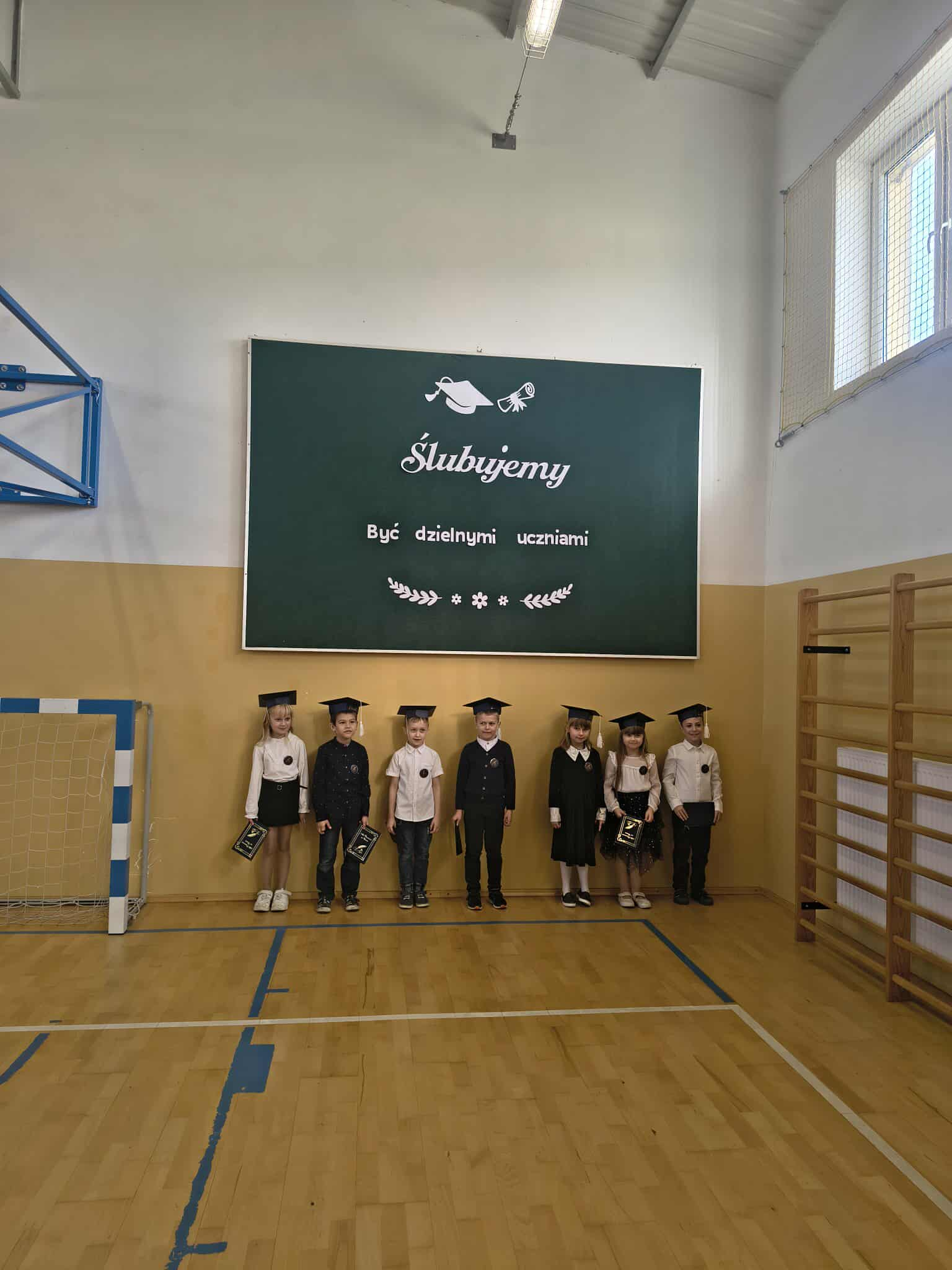 Klasa 1 stoi w szeregu pod tablicą „Ślubujemy”.