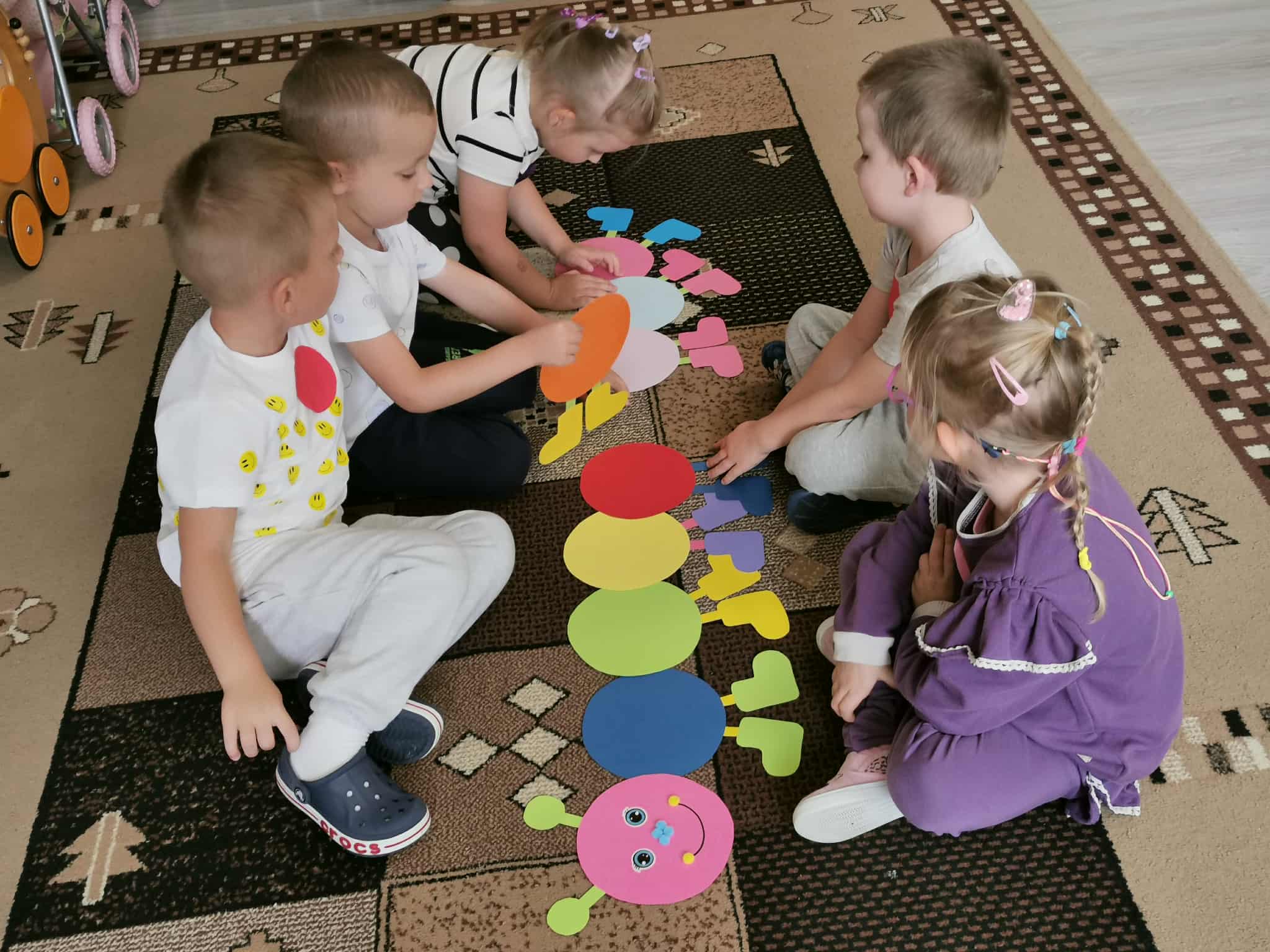 Pięcioro dzieci siedzi na dywanie i wspólnie układa gąsienicę z kolorowych papierowych kropek.