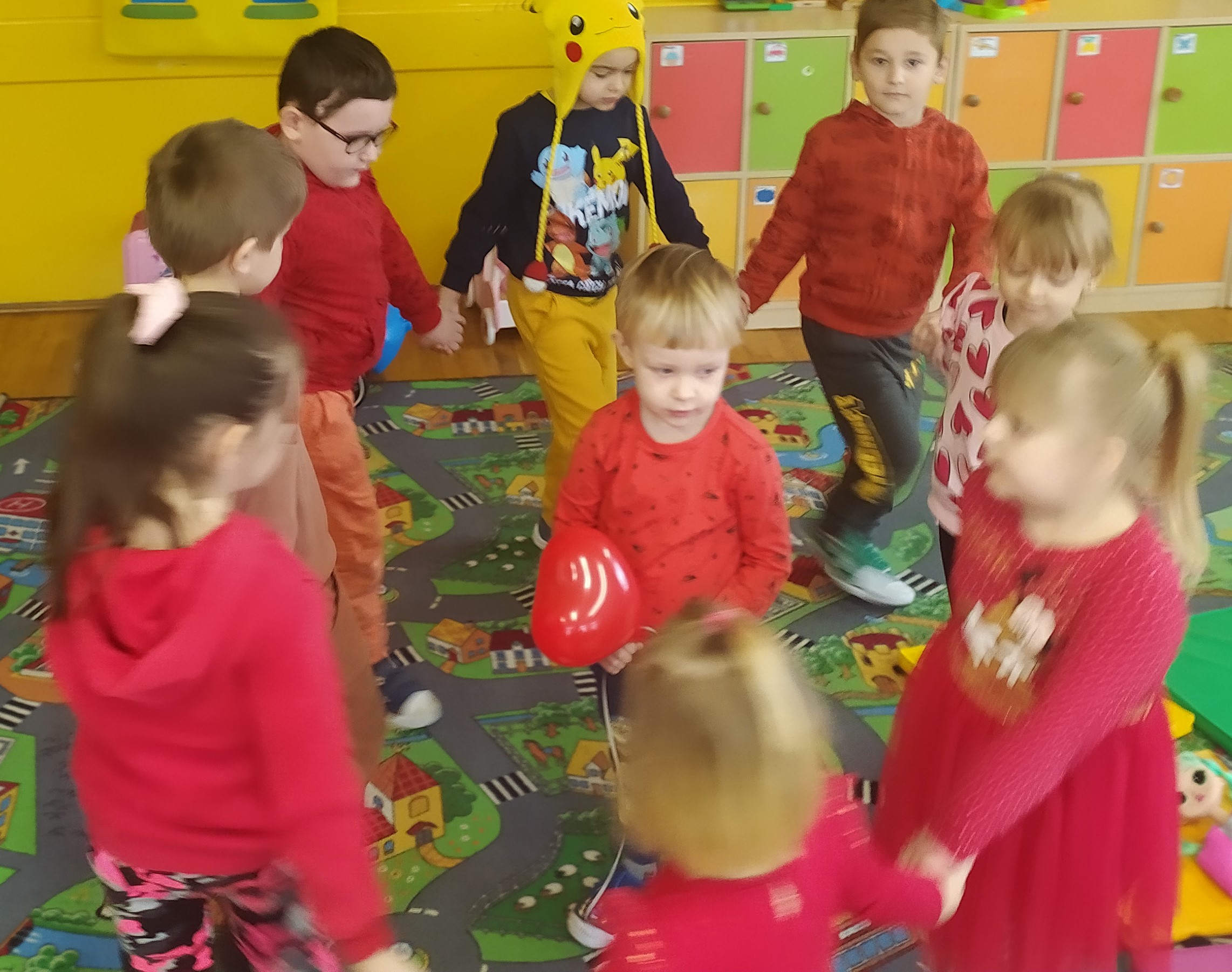 Dzieci tańczą w kole wokół chłopca trzymającego balonik w kształcie serca.