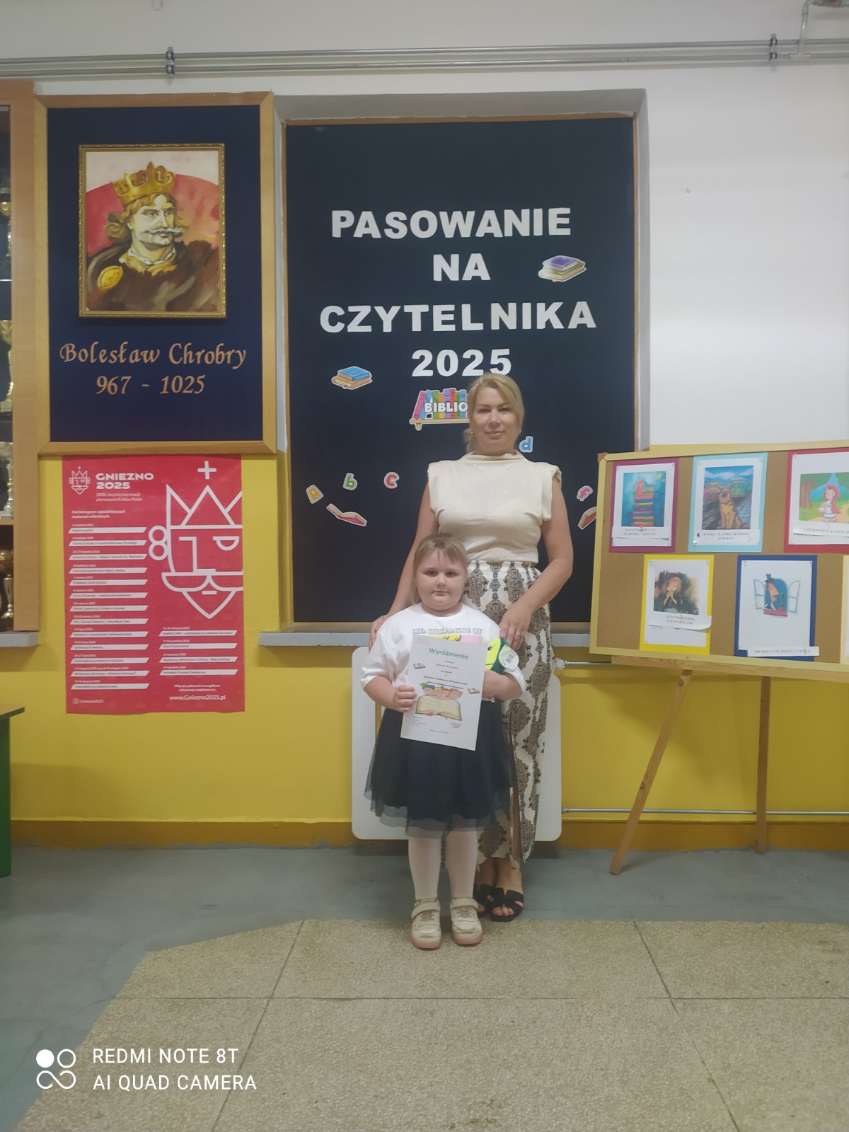 Dziewczynka stoi z nauczycielką na tle gazetki 