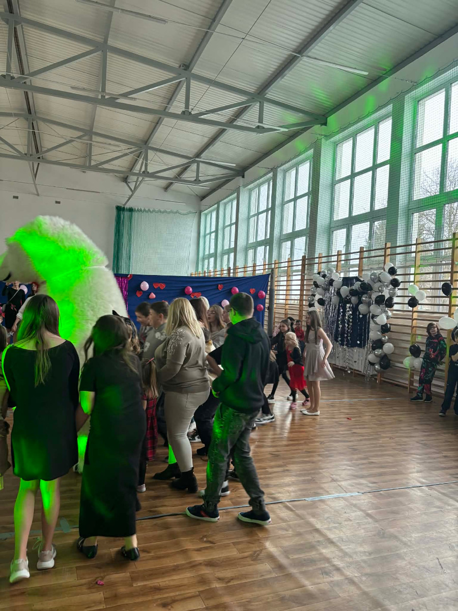 Dzieci wraz z uczniami biorą udział w zabawie choinkowej przy muzyce, a w środku tańczy biały miś.