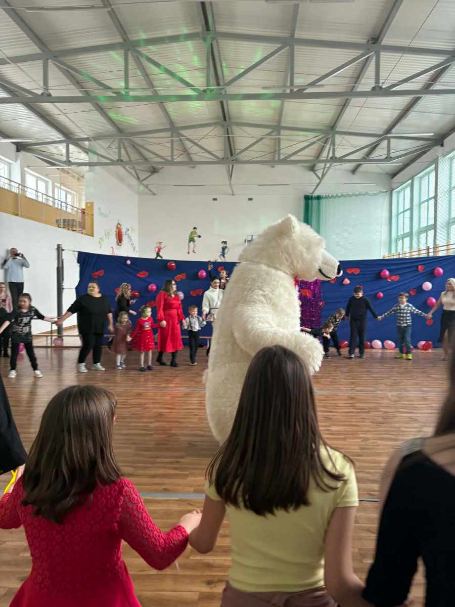 Dzieci wraz z uczniami biorą udział w zabawie choinkowej przy muzyce, a w środku tańczy biały miś.