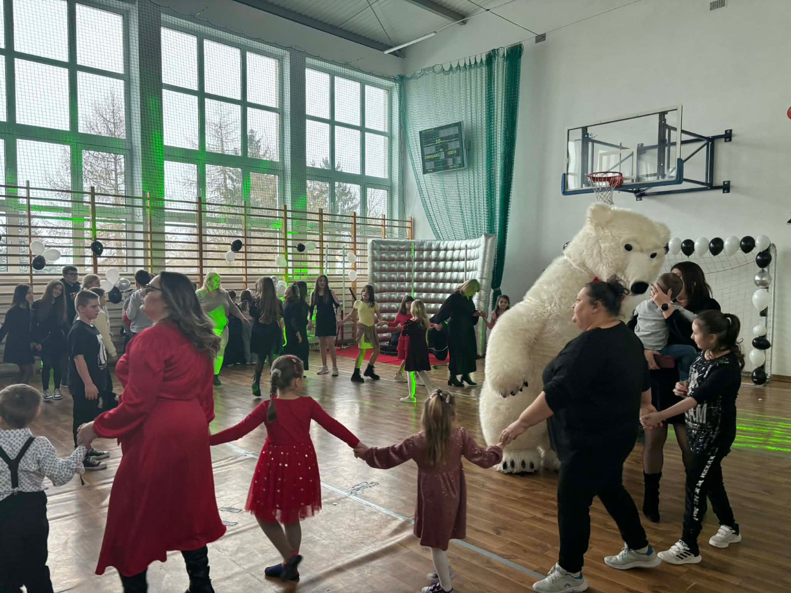 Dzieci wraz z uczniami biorą udział w zabawie choinkowej przy muzyce, a w środku tańczy biały miś.