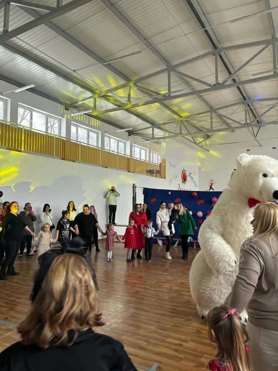 Dzieci wraz z uczniami biorą udział w zabawie choinkowej przy muzyce, a w środku tańczy biały miś.