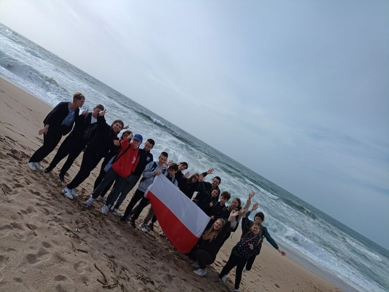 Zdjęcie przedstawia grupę uczniów, którzy znajdują się na plaży, trzymają flagę Polski. W tle widać ocean.