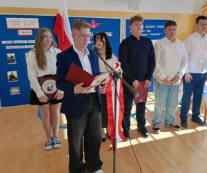 Jeden uczeń stoi przy mikrofonie, a pozostali z tyłu na tle dekoracji