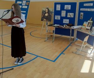 Uczennica czyta przy mikrofonie, a w tle dekoracja okolicznościowa