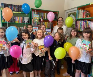 Zdjęcie przedstawia dzieci wraz z opiekunem na tle regałów z książkami.