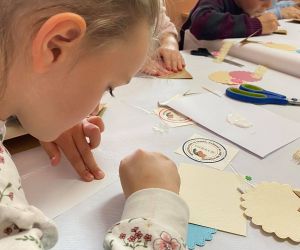 Dzieci podczas warsztatów organizowanych przez GOK, wewnątrz budynku, wykonują kartkę metoda scrapbookingu.