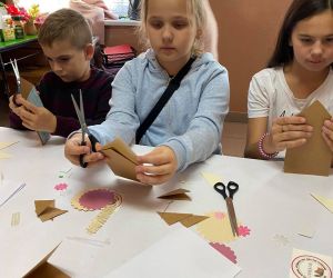 Dzieci podczas warsztatów organizowanych przez GOK, wewnątrz budynku, wykonują kartkę metoda scrapbookingu.