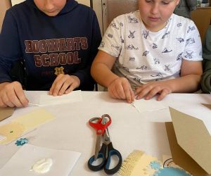 Dzieci podczas warsztatów organizowanych przez GOK, wewnątrz budynku, wykonują kartkę metoda scrapbookingu.