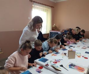 Dzieci znajdują sie wewnątrz budynku, siedzą przy stole i wykonują kartki okolicznościowe.