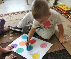  Chłopiec siedzi na dywanie i rysuje na kartce z kolorowymi kropkami, obok przygląda mu się koleżanka.