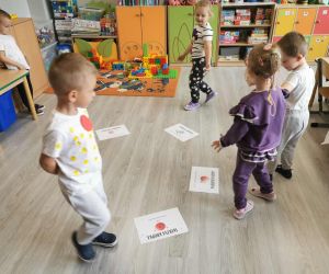 Dzieci bawią się w sali przedszkolnej. Na podłodze rozłożone są kartki z zadaniami związanymi z Dniem Kropki.