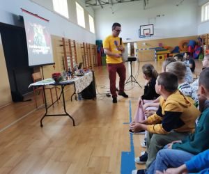 Podróżnik stoi blisko uczniów i opowiada, wskazując na jeden z przedmiotów ze stołu.