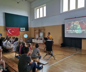 Podróżnik siedzi obok stołu z pamiątkami, prowadzi prezentację wyświetlaną na ekranie. Uczniowie aktywnie zgłaszają się do odpowiedzi.