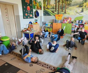 Dzieci z oddziału przedszkolnego leżą na podłodze i wykonują ćwiczenia gimnastyczne z uniesionymi nogami.