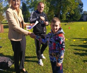 Dyrektor szkoły i nauczyciel wychowania fizycznego wręczają dyplom i medal za pierwsze miejsce uczniowi klasy drugiej