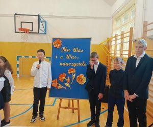 Czterech uczniów na tle tablicy dekoracyjnej, jeden z nich przemawia do mikrofonu