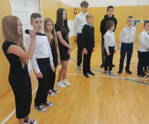 Uczniowie stoją półkolem, jedna z uczennic śpiewa do mikrofonu