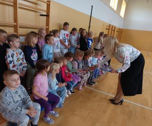 Przedstawicielka Banku Pekao S.A. wręcza uczniom kolorowanki i ulotki podczas spotkania w sali gimnastycznej. Dzieci siedzą i stoją w rzędzie, z zainteresowaniem przyjmując materiały.