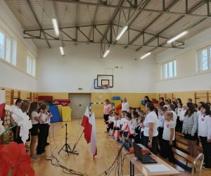 Uczniowie i nauczyciele stoją w sali gimnastycznej podczas akademii z okazji Święta Niepodległości.