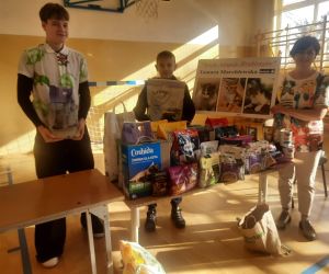 Pani Tamara Marchlewska i dwaj uczniowie klasy siódmej przy stoliku z zebranymi darami prezentują wizytówkę 