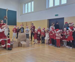 Mikołaj rozmawia z najmłodszymi dziećmi ustawionymi w szeregu na sali gimnastycznej.