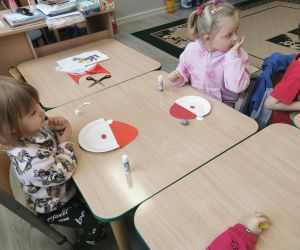 Dzieci przy stoliku przygotowują elementy papierowej czapki do pracy plastycznej.