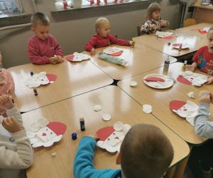 Przedszkolaki siedzą przy stolikach i wykonują prace plastyczne przedstawiające Mikołaja z papierowych talerzyków i płatków kosmetycznych.