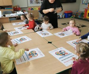 Dzieci z oddziału przedszkolnego siedzą przy stolikach w sali i wykonują kalendarze, pracując z kartkami papieru pod opieką nauczycielki.