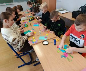 Grupa uczniów pracuje przy jednym stole, wycinając i przyklejając elementy dekoracyjne na kartkach podczas warsztatów plastycznych.