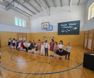 Dzieci 3-, 4-, 5- i 6-letnie ustawione w jednym szeregu na środku sali gimnastycznej podczas uroczystości z okazji Dnia Babci i Dziadka. W tle widoczna dekoracja okolicznościowa.