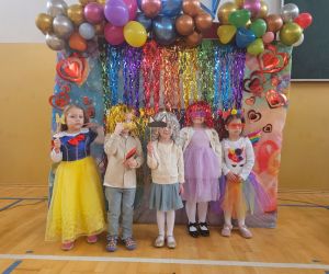 Pięcioro dzieci w kolorowych strojach i przebraniach pozujących przy dekoracyjnej ściance z balonami i tęczowymi wstążkami podczas szkolnej zabawy choinkowej.