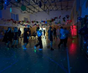 Uczniowie tańczący w kole na sali gimnastycznej przy przyciemnionym świetle i kolorowych efektach świetlnych, sufit udekorowany balonami.