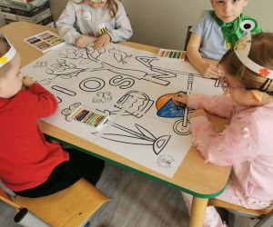 Dzieci siedzą przy stoliku i kolorują dużą wspólną pracę plastyczną związaną z wiosną, korzystając z kredek.