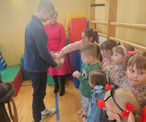 Starszy uczeń losuje elementy z pojemnika, a młodsze dzieci ustawione w kolejce obserwują i czekają na swoją kolej podczas zabawy.